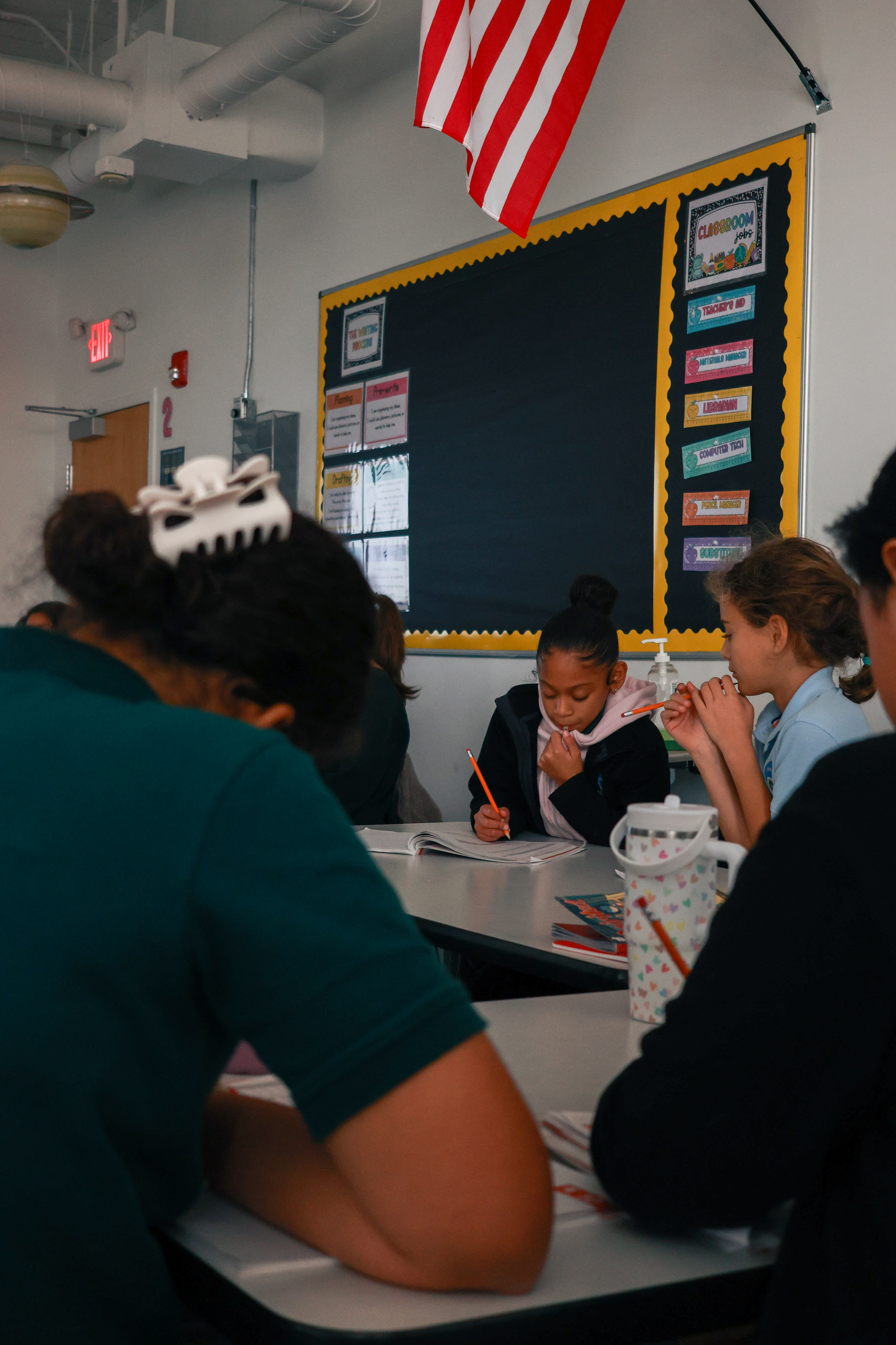 Photo of a student writing on a workbook, viewed over the shoulders of other students who are also working.